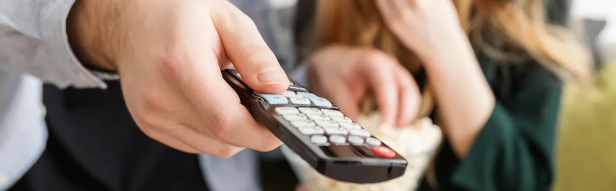 Close-up of a hand holding a TV remote, with a person in the background holding a bowl of popcorn